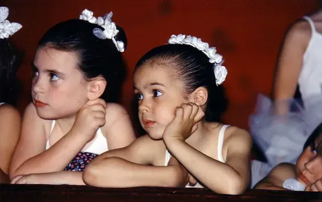 Dos jóvenes bailarinas con leotardos blancos y diademas florales apoyan la barbilla en una cornisa oscura, mirando fijamente hacia la izquierda.