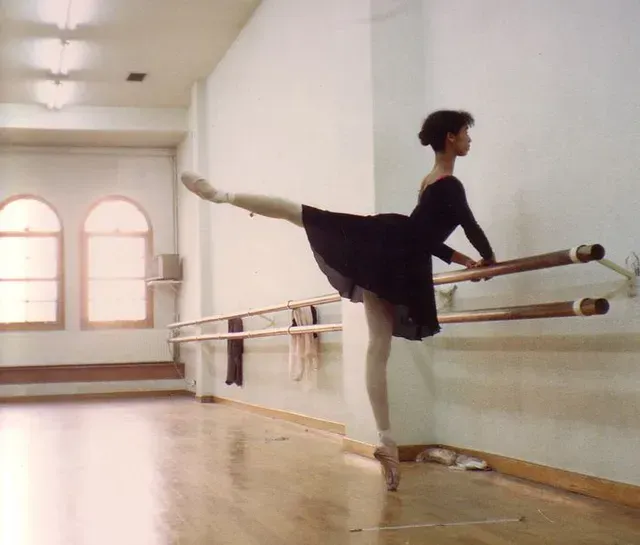 Bailarina con leotardo negro, manteniendo el equilibrio en punta con la pierna extendida en la barra del estudio de danza.