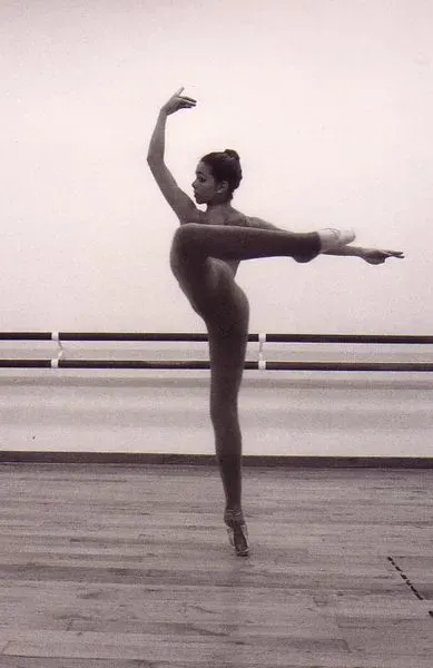 Bailarina en puntas, manteniendo el equilibrio con la pierna elevada y el brazo extendido. Escenario de estudio con barra.