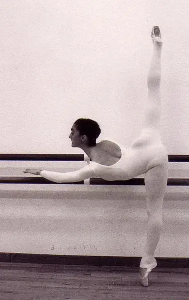 Bailarina de ballet con leotardo blanco, realizando una apertura de piernas alta, sujeta a la barra.