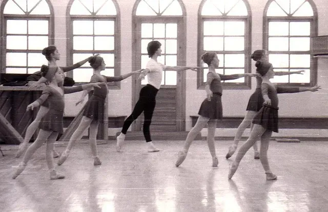 Clase de ballet en un estudio de danza. Las bailarinas, con tutús, practican movimientos de brazos dirigidas por una instructora.