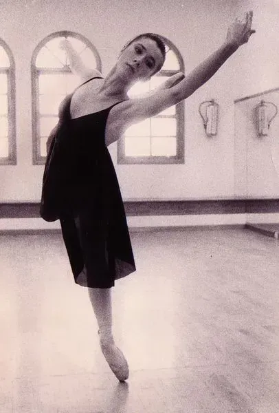 Bailarina con vestido negro, en puntas, con los brazos extendidos, en un estudio de danza.