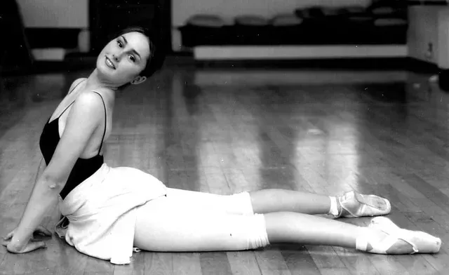 Bailarina con leotardo negro y pantalones cortos blancos, arqueada sobre un suelo de madera, con zapatillas de punta, sonriendo.