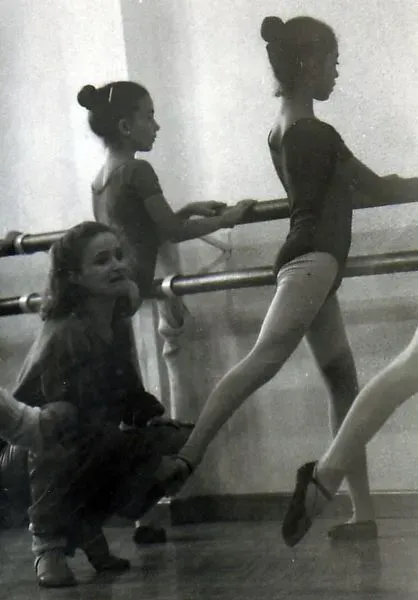 Clase de ballet: Jóvenes estudiantes en la barra, estirando las piernas. Una profesora les ayuda.