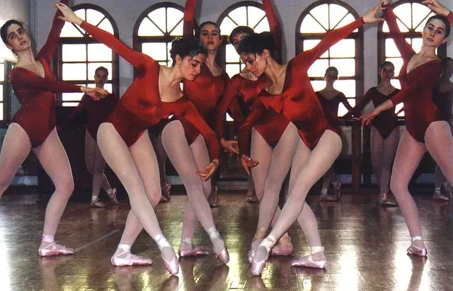 Bailarinas de ballet con leotardos rojos realizan una rutina en un estudio de danza con grandes ventanales.