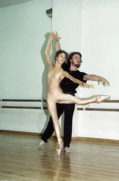 Una bailarina con un traje dorado es alzada por un hombre en un estudio de ballet, con las piernas extendidas en actitud de baile.