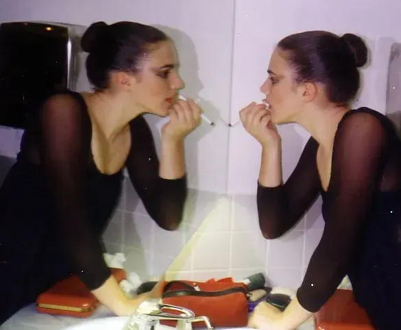 Mujer con leotardo negro aplicándose maquillaje frente al espejo de un baño; su reflejo es visible.