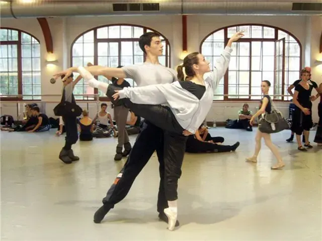 Bailarinas de ballet en un estudio, una alzada por otra. Interior, otras personas observando.