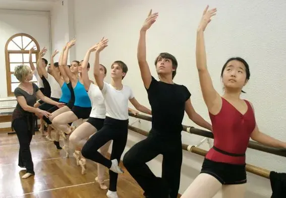 Clase de ballet en la barra. Alumnas con ropa deportiva, brazos levantados, practicando bajo la supervisión de la instructora. Estudio luminoso.