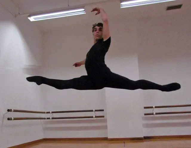 Bailarín de ballet masculino vestido de negro realizando un salto con piernas abiertas en un estudio de danza.