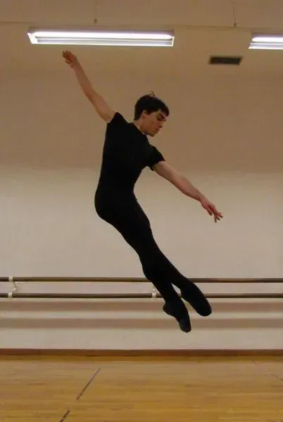 Bailarín de ballet masculino saltando en el aire, con los brazos extendidos, vestido con leotardo y mallas negras en un estudio de danza.