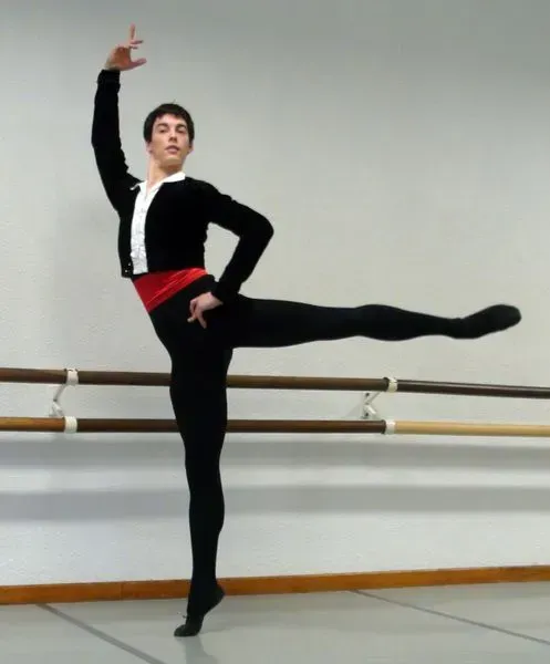 Bailarina de ballet con mallas negras, chaqueta y fajín rojo, en pose de arabesco, con el brazo levantado. Fondo de estudio de ballet.