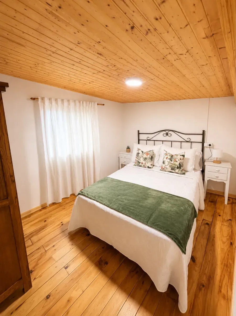 Habitación acogedora con suelo y techo de madera, cama blanca, manta verde y cortina transparente en la ventana.