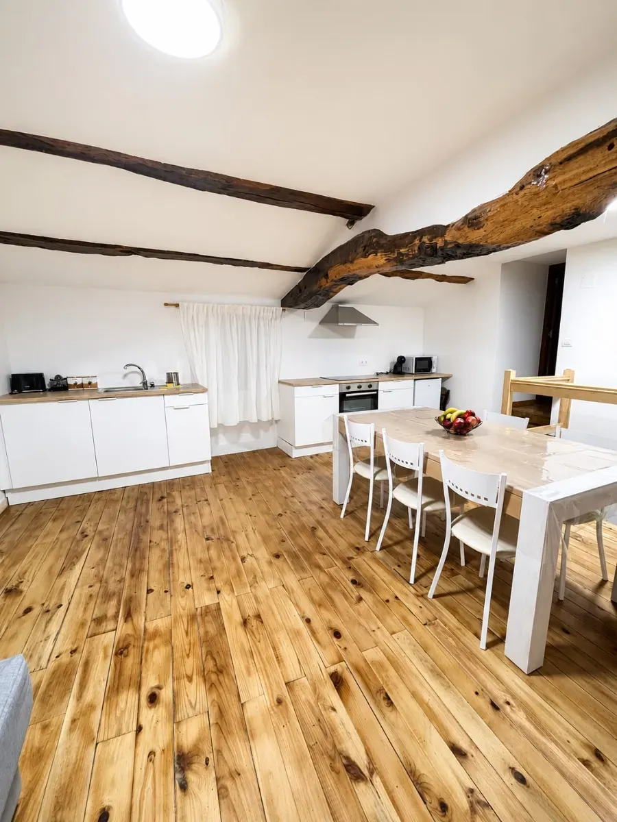 Comedor luminoso de color blanco con vigas de madera rústica, suelo de madera, armarios blancos y una mesa con sillas.