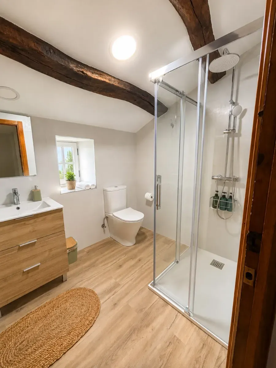 Baño moderno con ducha de cristal, lavabo de madera, inodoro y vigas vistas.