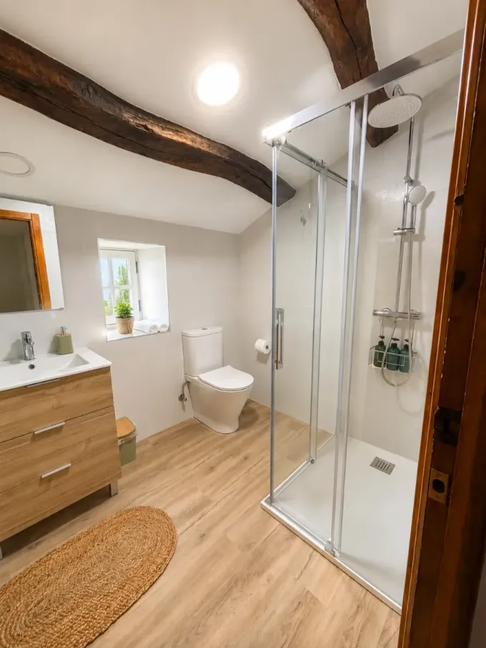Baño luminoso con vigas de madera, ducha de cristal, lavabo, inodoro y alfombra de mimbre.