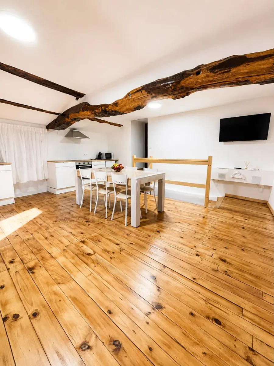 Comedor luminoso en el ático con suelo de madera, viga vista, mesa blanca, sillas y televisor montado en la pared.