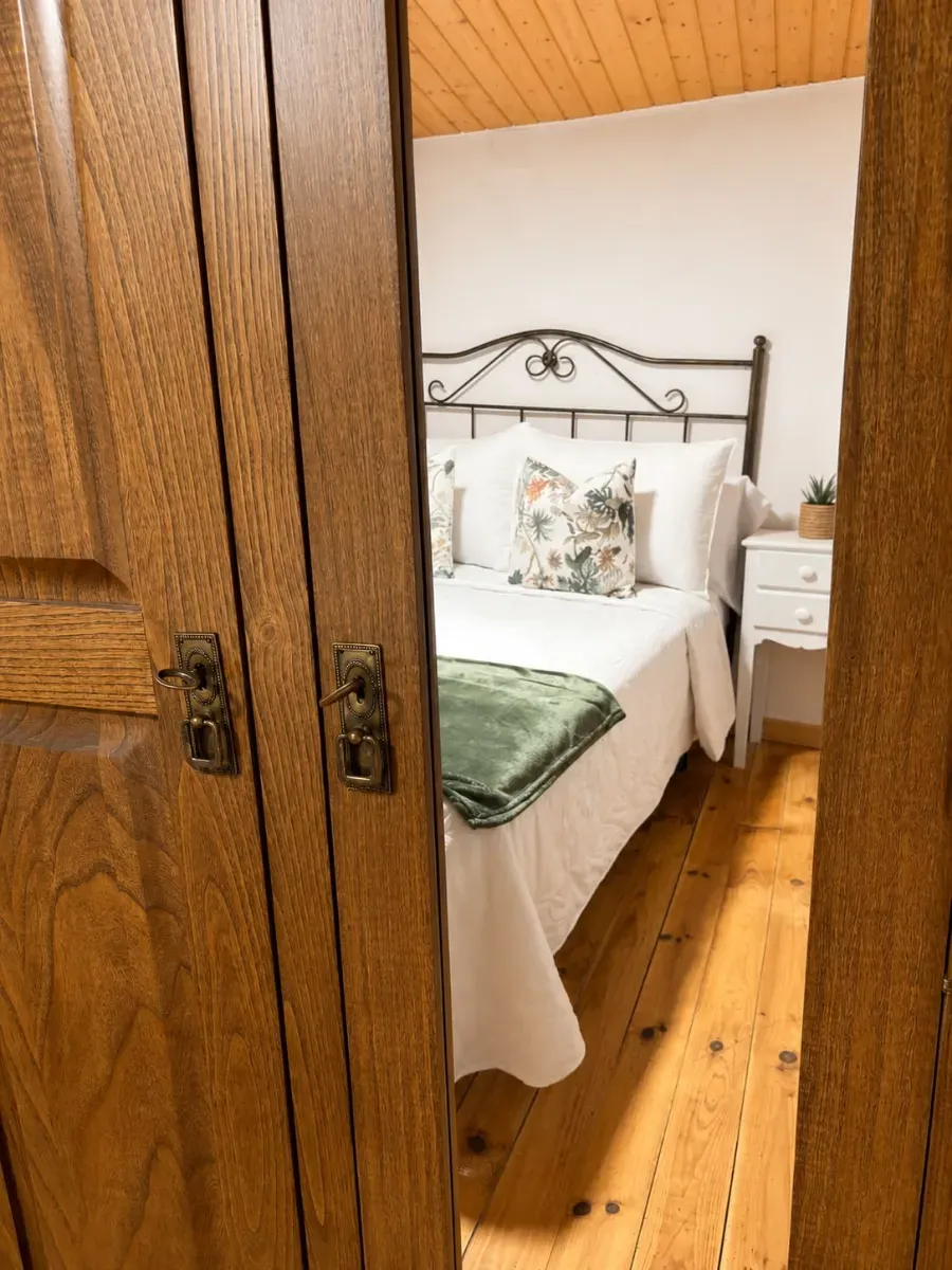 Pequeña habitación vista a través de las puertas de un armario de madera, con una cama blanca, una manta verde y una mesita de noche.