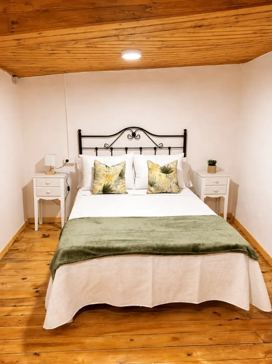 Acogedora habitación con ropa de cama blanca, manta verde, cabecero de metal negro, techo y suelo de madera y mesitas de noche a juego.