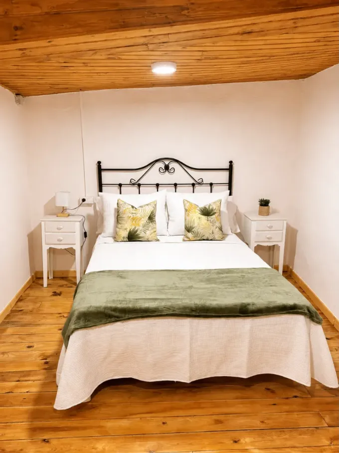 Habitación acogedora con cama, mesitas de noche blancas, manta verde y techo y suelo de madera.