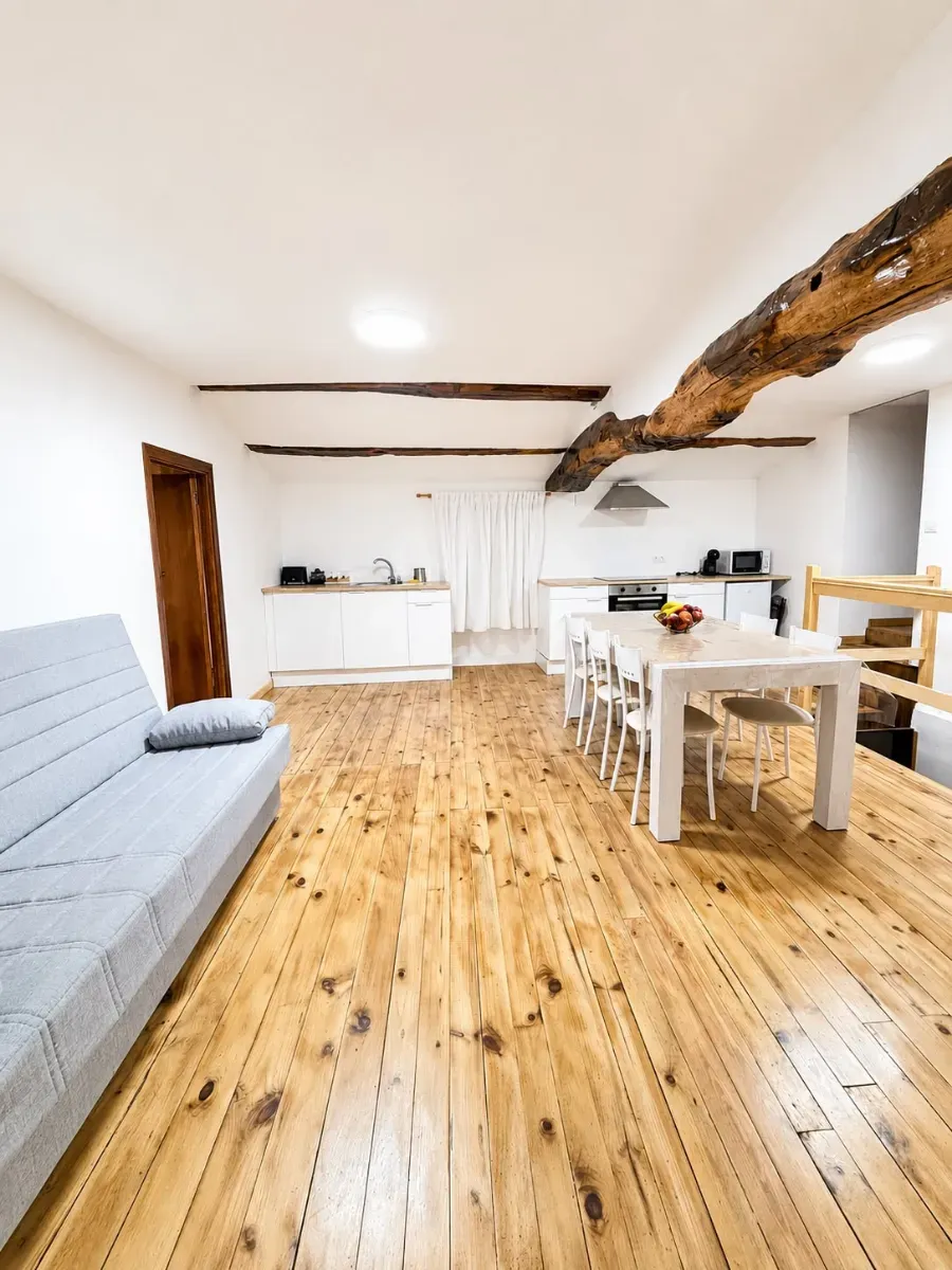 Salón luminoso y diáfano con suelos de madera, paredes blancas, sofá gris, mesa de comedor y viga de techo a la vista.