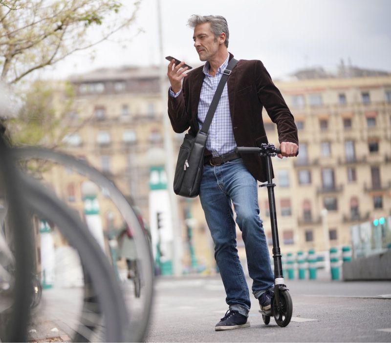 Ruskeaan bleiseriin ja farkkuihin pukeutunut henkilö seisoo mustan sähköpotkulaudan kanssa puhuen älypuhelimeen kaupungissa.