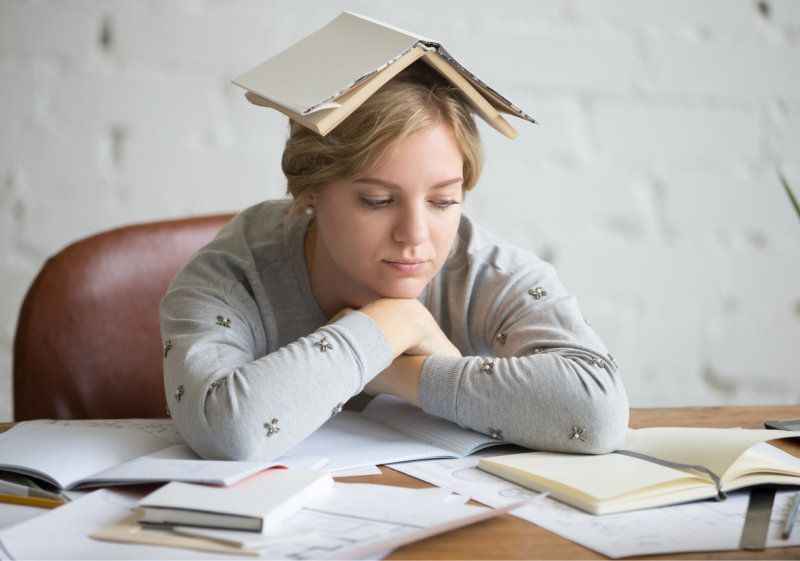 Opiskelija istuu pulpetin ääressä avoin kirja päänsä päällä, papereiden ympäröimänä, väsyneenä ja hämmentyneenä.