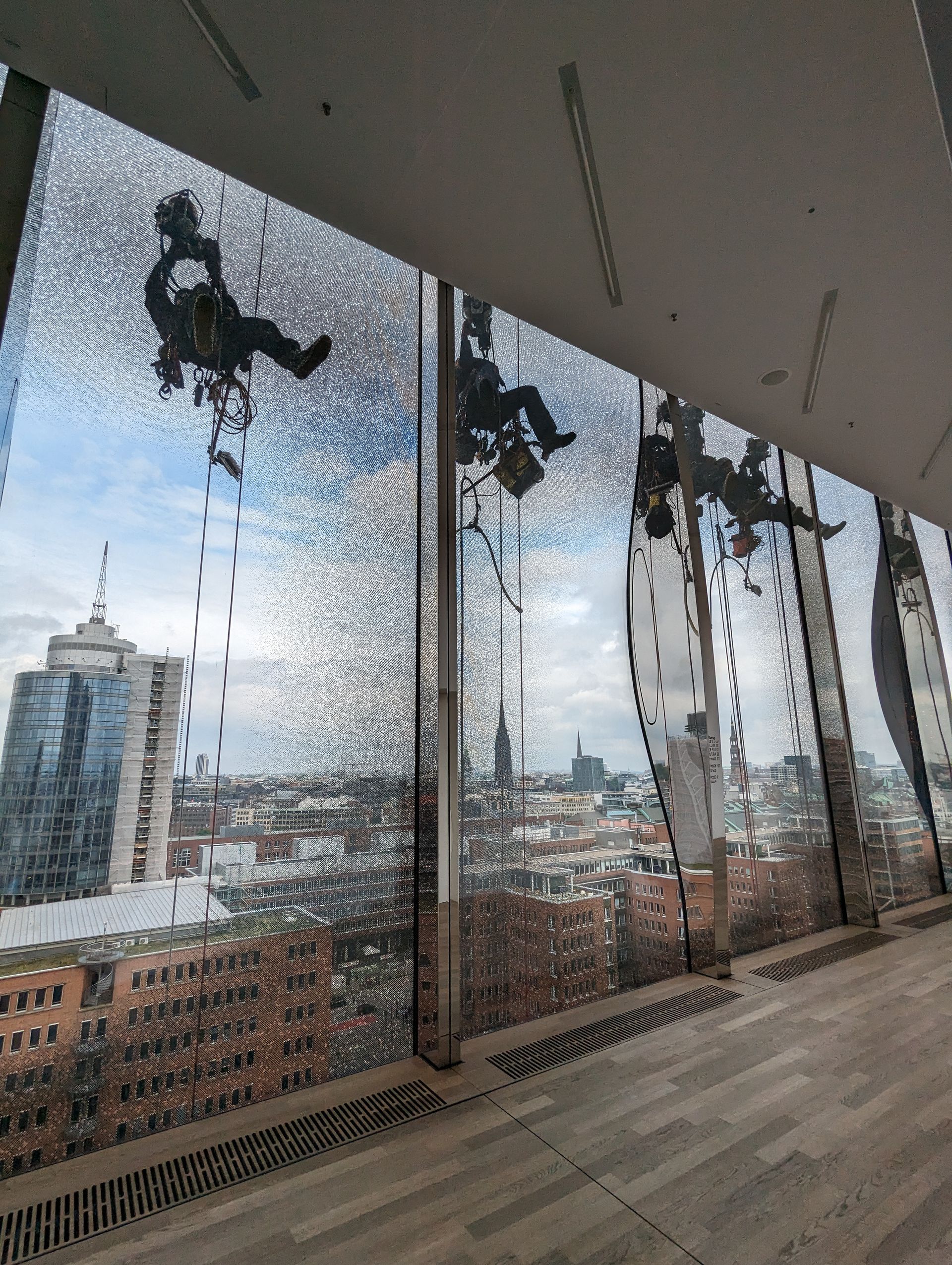 Arbeiter hängen an der Außenseite eines Glasgebäudes und putzen die Fenster. Im Hintergrund ist die Stadtkulisse Hamburgs zu sehen.