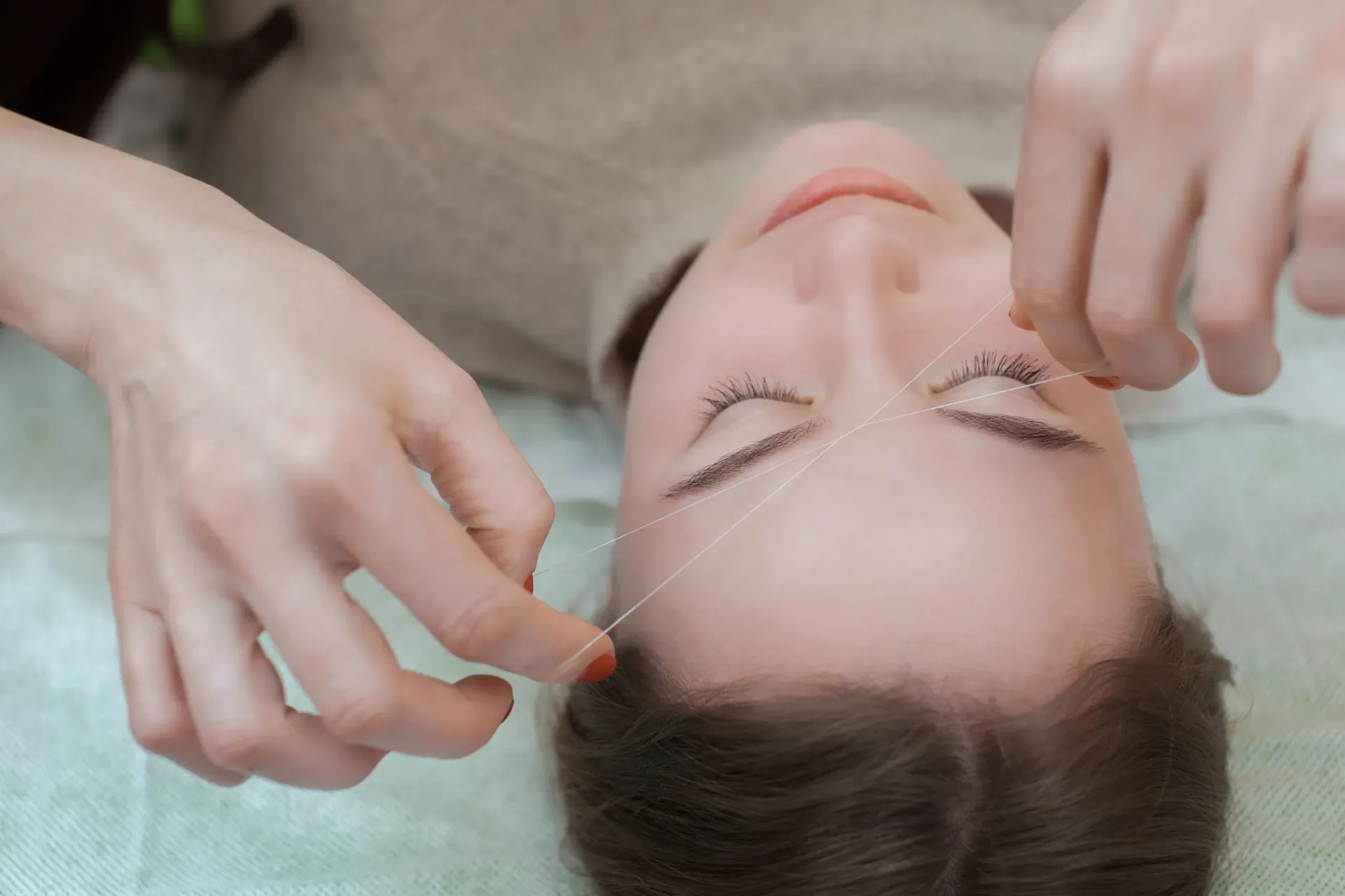 Una persona realiza la depilación de cejas con hilo a una clienta tumbada, utilizando un hilo blanco para eliminar el vello.