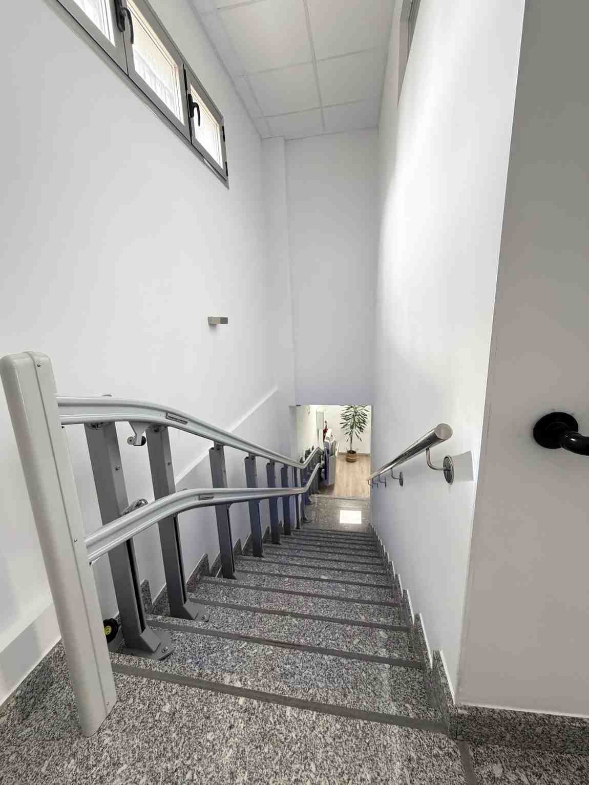 Escalera con peldaños de piedra, paredes blancas y una barandilla plateada para silla salvaescaleras en el lado izquierdo.