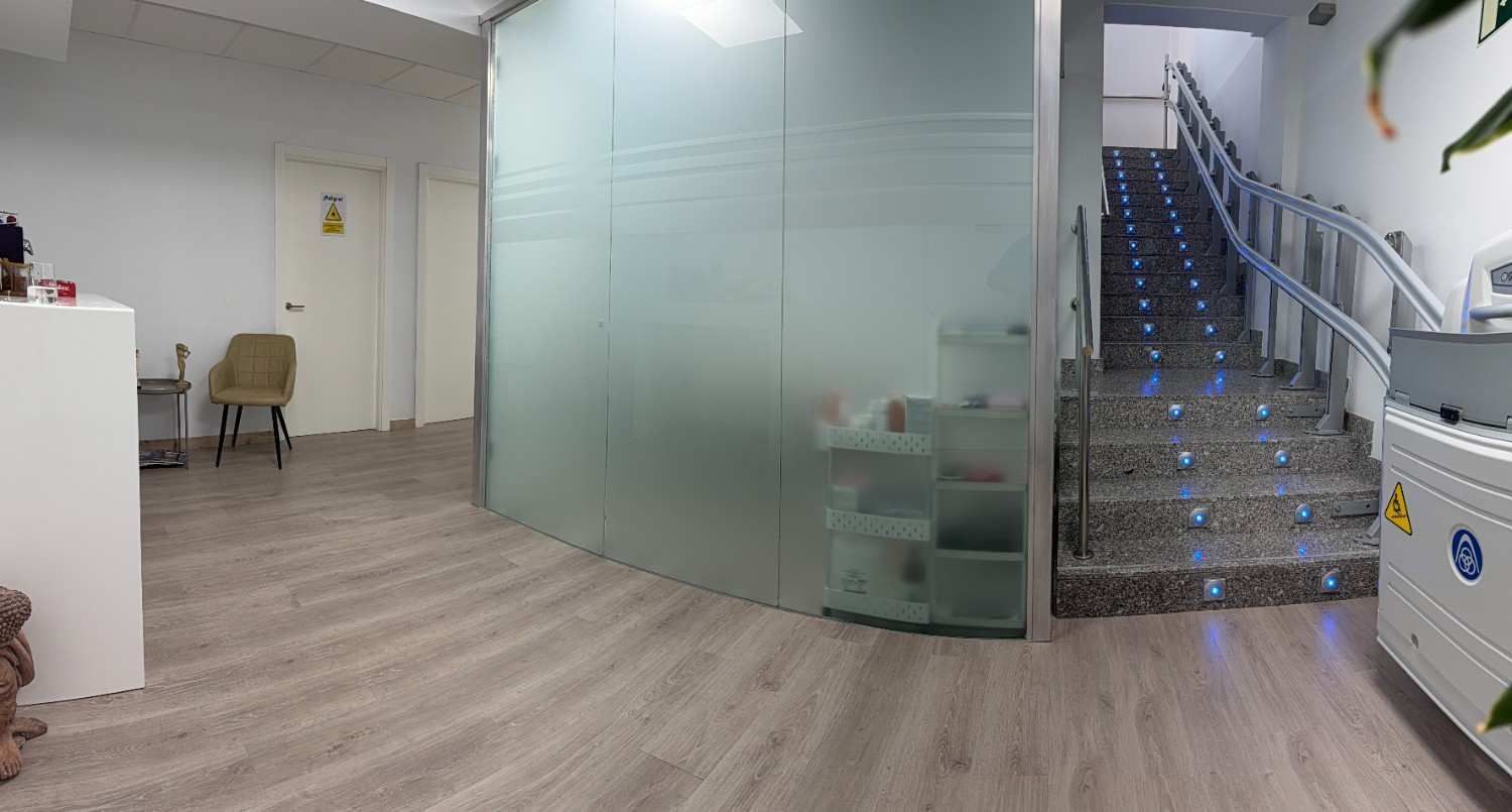 Un pasillo de oficina con suelo de madera clara, una mampara de vidrio esmerilado, una silla de color beige.