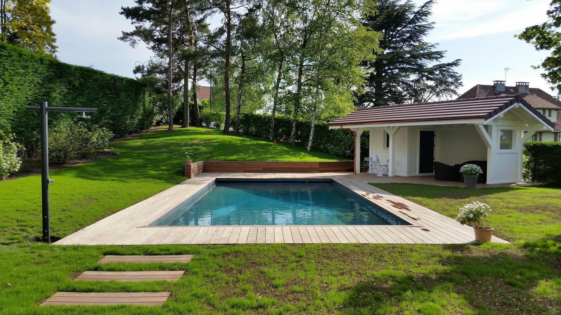 Piscine dans le jardin avec une terrasse en pierre, à côté d’un petit pool house entouré d’une pelouse.