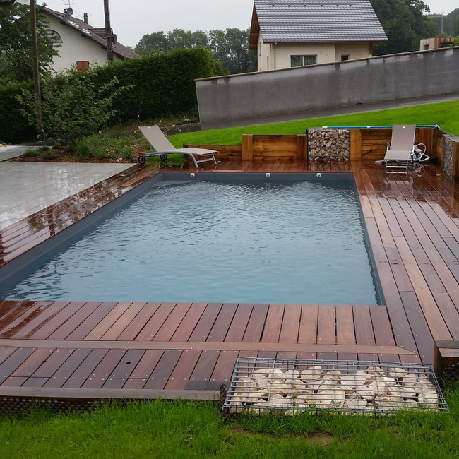 Piscine de jardin détrempée par la pluie, avec une terrasse en bois, des chaises longues et une pelouse.