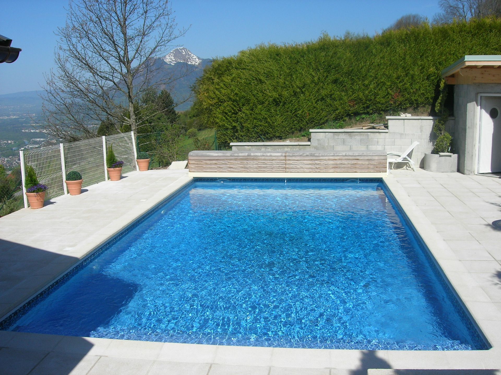 Piscine rectangulaire bleue à côté d’une haie et d’une terrasse, avec des montagnes.