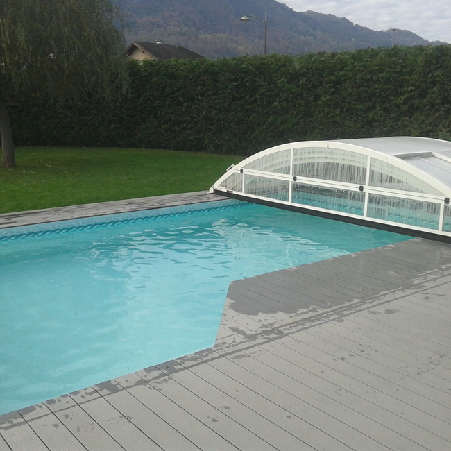 Piscine dans un jardin avec une terrasse une haie et un abri de piscine.