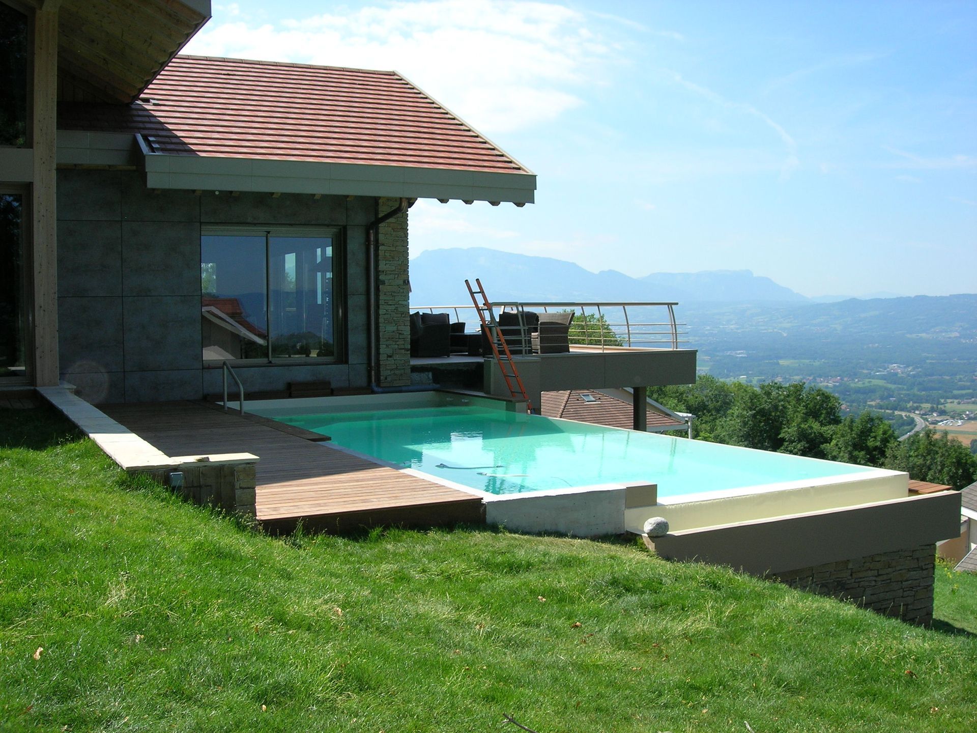 Maison moderne avec une piscine à débordement turquoise donnant sur une vallée et des montagnes au loin.