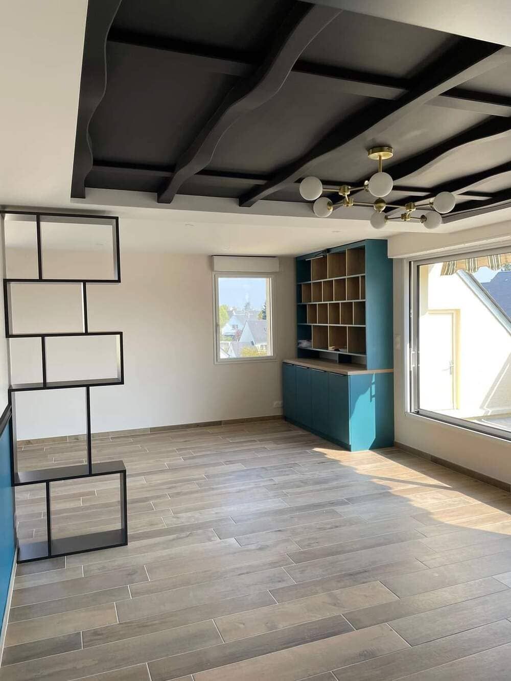 Une pièce avec un parquet clair, un plafond à caissons noirs, une cloison métallique et des étagères encastrées bleues près d'une fenêtre.