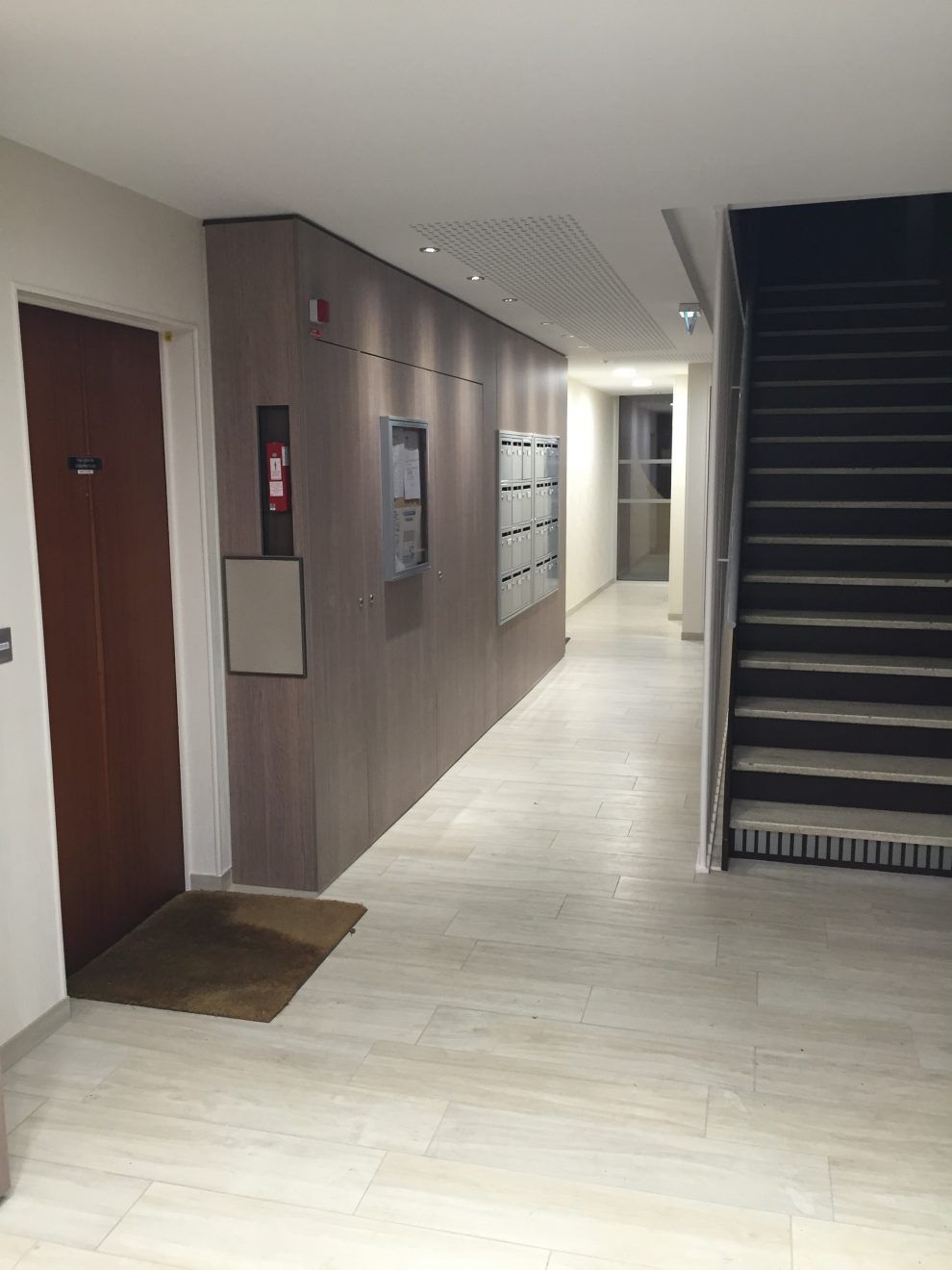 L'entrée d'un immeuble d'appartements moderne, avec un mur en bois foncé, des boîtes aux lettres en métal et un escalier menant à un couloir.