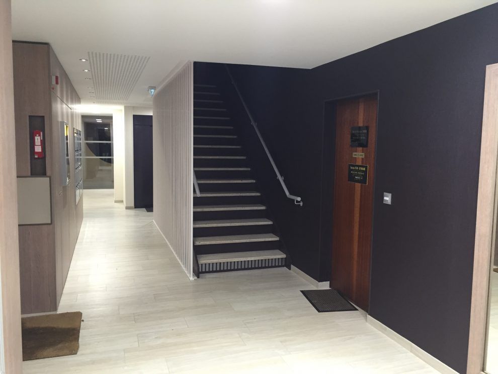 Un couloir intérieur au sol carrelé clair mène à un escalier en bois foncé, entre un mur blanc et un mur d'accent sombre.