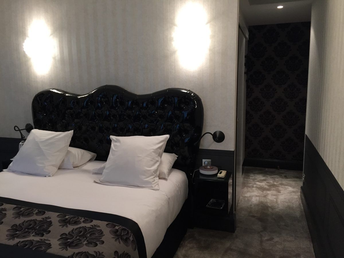 Une chambre avec une tête de lit capitonnée noire, du linge de lit blanc avec un chemin de lit à motifs foncés et un éclairage mural doux.