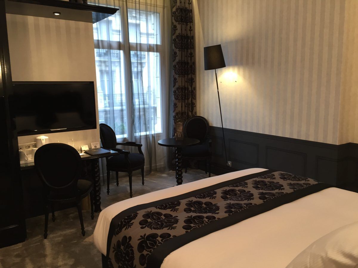 Une chambre d'hôtel avec un lit recouvert d'un chemin de table gris à motifs, deux chaises noires, une table et un lampadaire près d'une fenêtre.