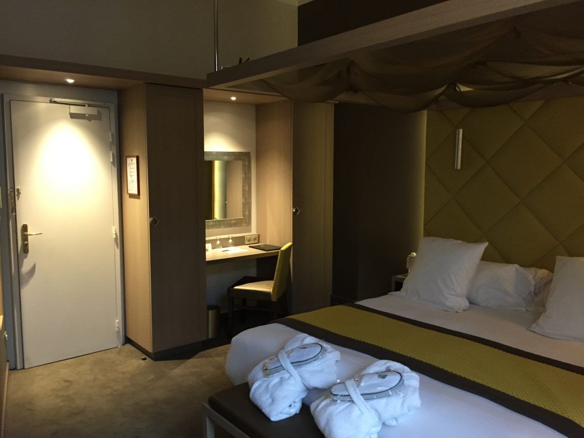Une chambre d'hôtel avec un lit fait, deux peignoirs blancs au pied du lit, un bureau avec un miroir et une porte aux tons chauds sur la gauche.