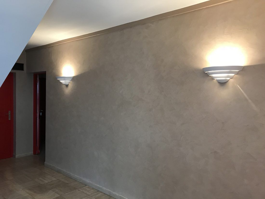 Un mur taupe avec deux appliques murales blanches à plusieurs niveaux éclairant un couloir faiblement éclairé près d'une porte rouge.
