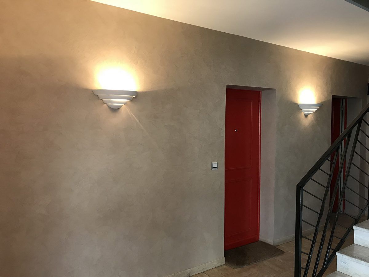 Un mur beige avec deux appliques murales encadrant une porte rouge vif, à côté d'un escalier avec une rampe en métal noir.