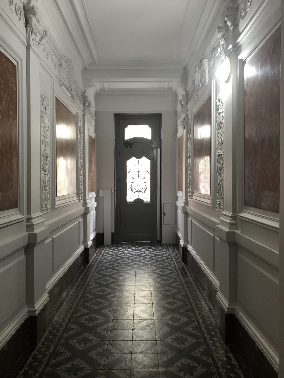 Un couloir étroit et faiblement éclairé, aux murs lambrissés blancs, orné de moulures décoratives et d'un sol carrelé à motifs sombres, menant à une porte.