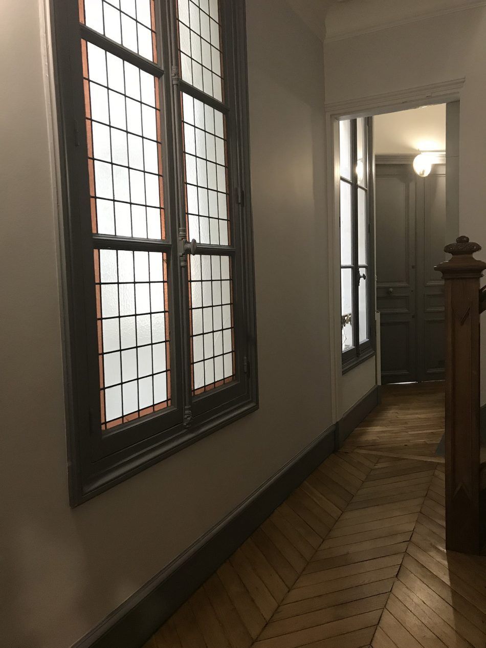 Un couloir lumineux avec une fenêtre en verre plombé sur la gauche, un parquet à chevrons et une porte ouverte donnant sur une autre pièce.