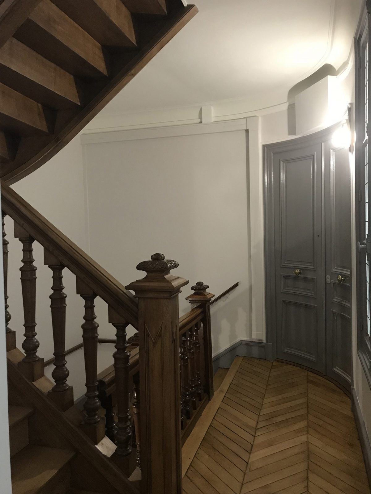 Un escalier intérieur en bois avec une rampe, à côté d'une porte à panneaux gris et d'un sol en bois clair à chevrons.