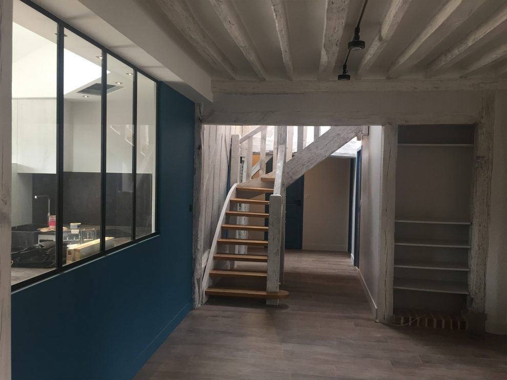 Vue intérieure d'une pièce avec un mur bleu, une cloison en verre, un escalier en bois et des étagères ouvertes.
