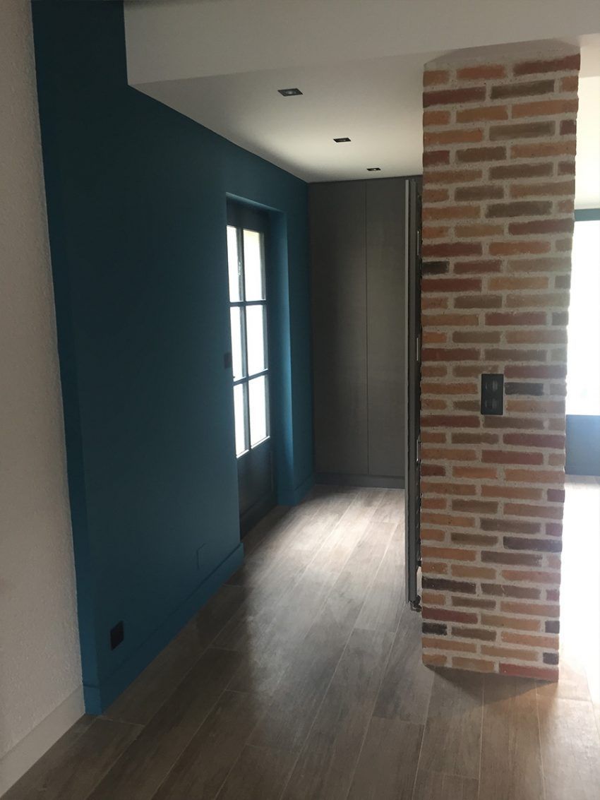 Le couloir présente un mur peint en bleu sarcelle foncé, un pilier en briques, un parquet clair et une porte vitrée.