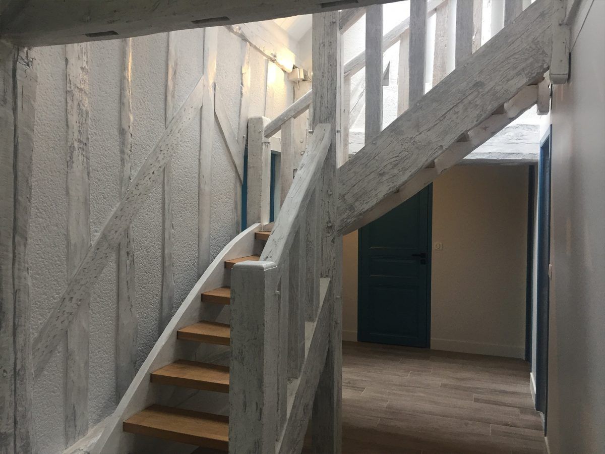Un escalier rustique en bois aux poutres peintes en blanc mène à l'étage, longeant un mur texturé et une porte bleu foncé.